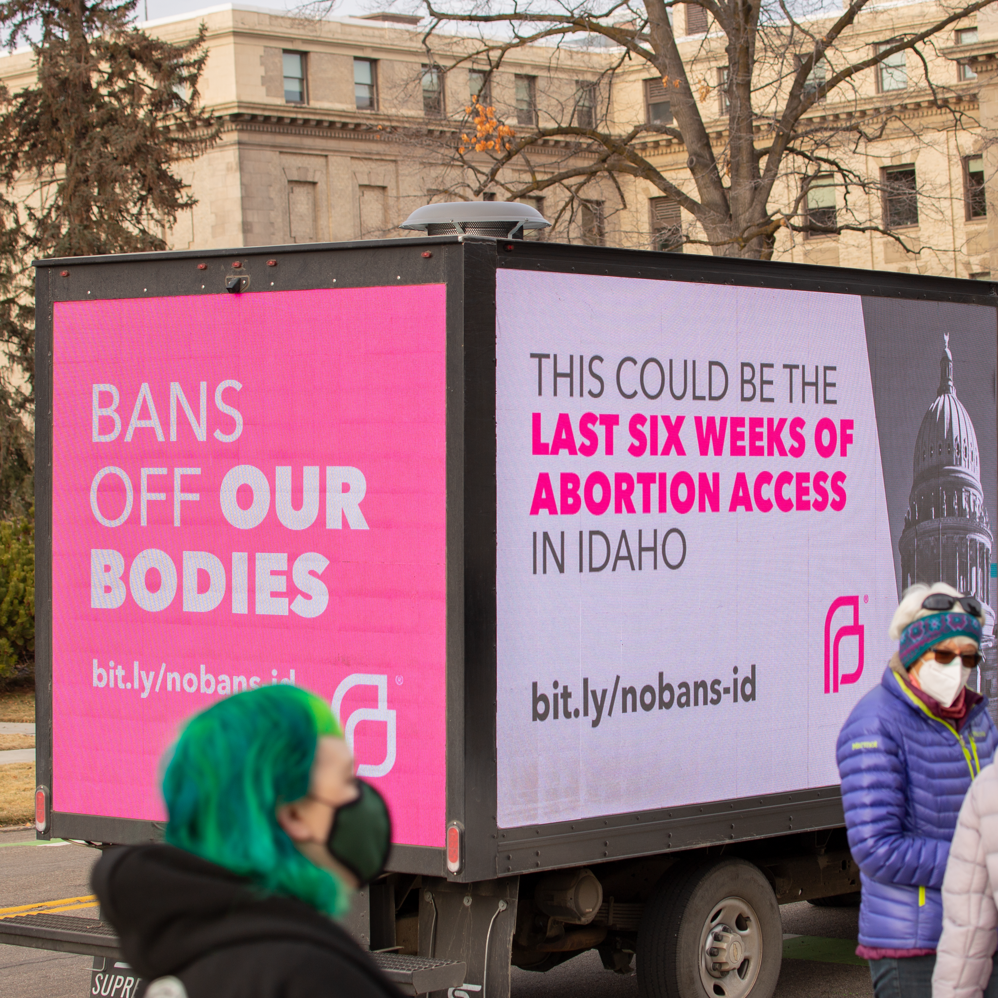 Billboard truck with protesters standing around it. One side is pink and reads Bans Off Our Bodies, and the other is white and reads This could be the last 6 weeks of abortion access in Idaho.